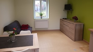 A compact living space with a dining area, a sofa, a TV mounted on the wall, and a window with a plant.