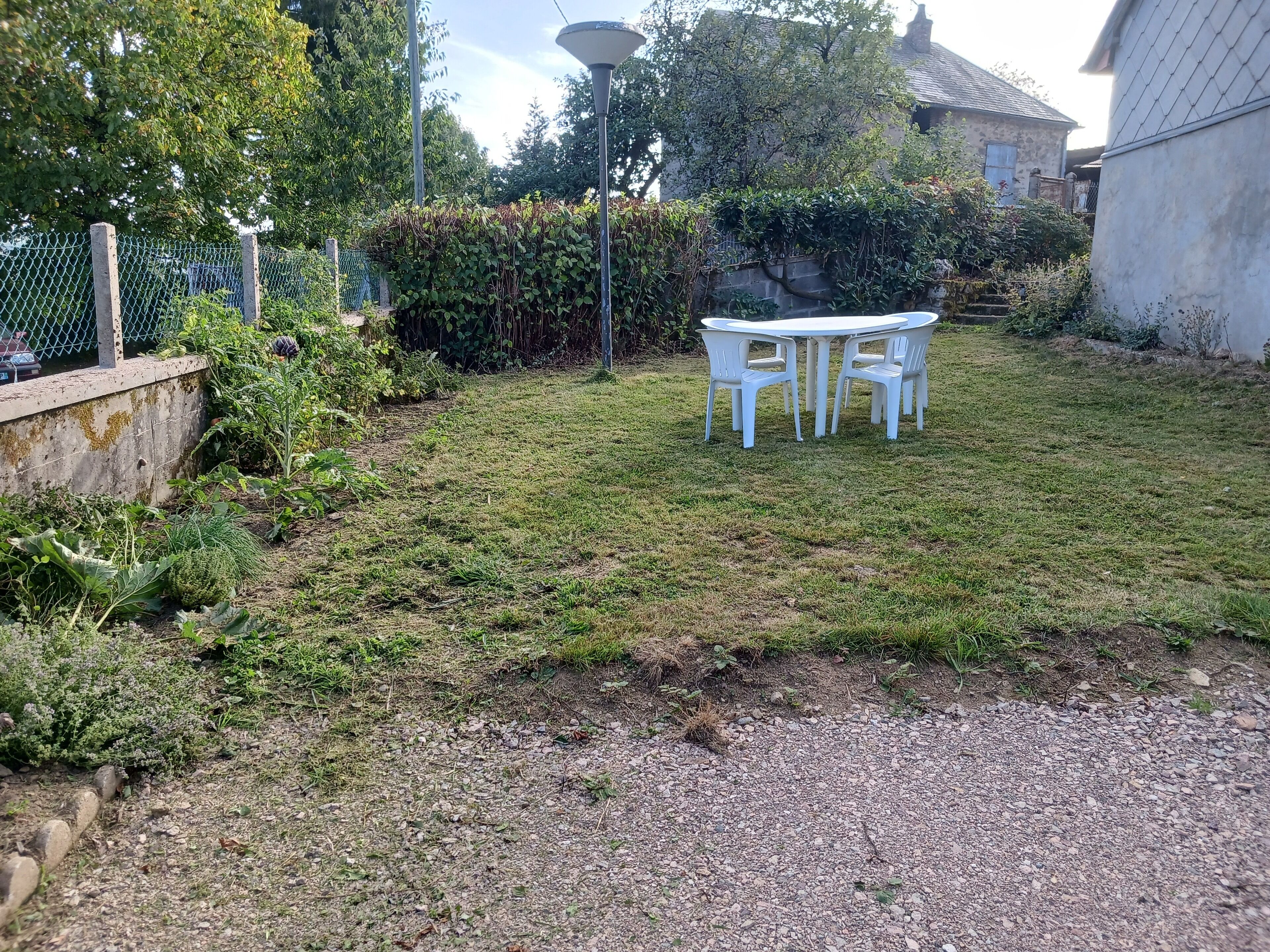A grassy outdoor area with a white table and chairs, surrounded by greenery and a stone wall.