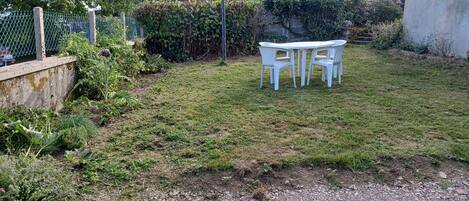 A grassy outdoor area with a white table and chairs, surrounded by greenery and a stone wall.
