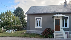 A two-story house with a grey exterior, a front door, and a small porch with steps. There is a table and chairs outside, and a hedge near the house.