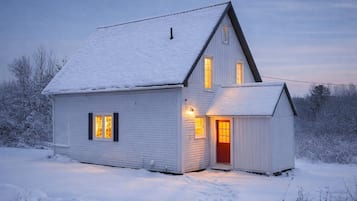 Une maison recouverte de neige, avec une porte rouge et deux fenêtres illuminées.