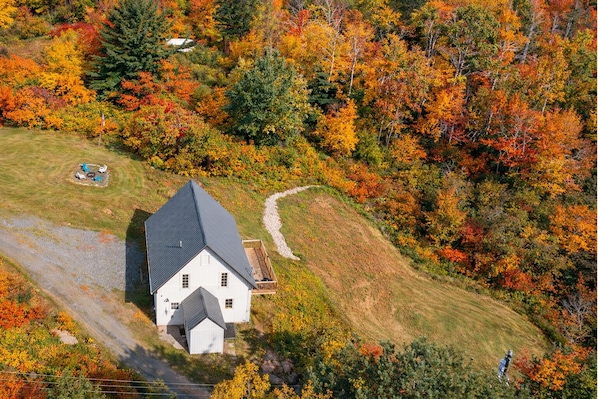 Une maison blanche avec un toit foncé, entourée d’une cour verdoyante et d’un chemin de terre menant à une terrasse en bois.
