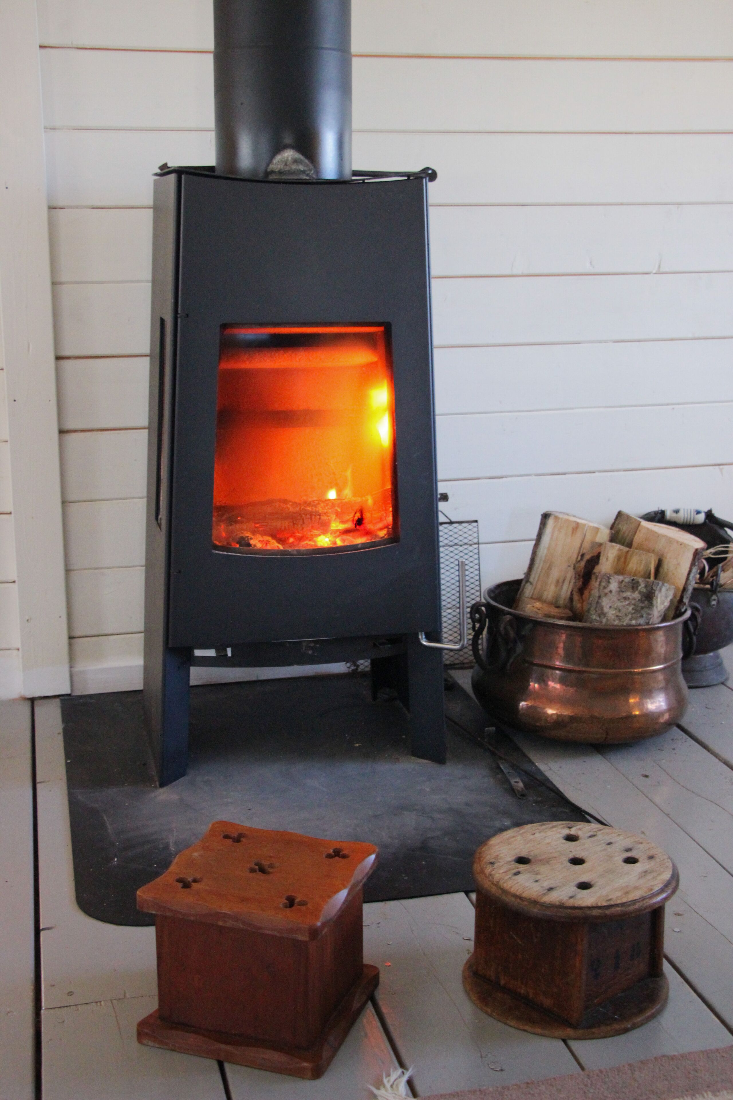 Un poêle à bois dont on peut voir les flammes, une marmite en métal remplie de bûches et un petit tabouret en bois.