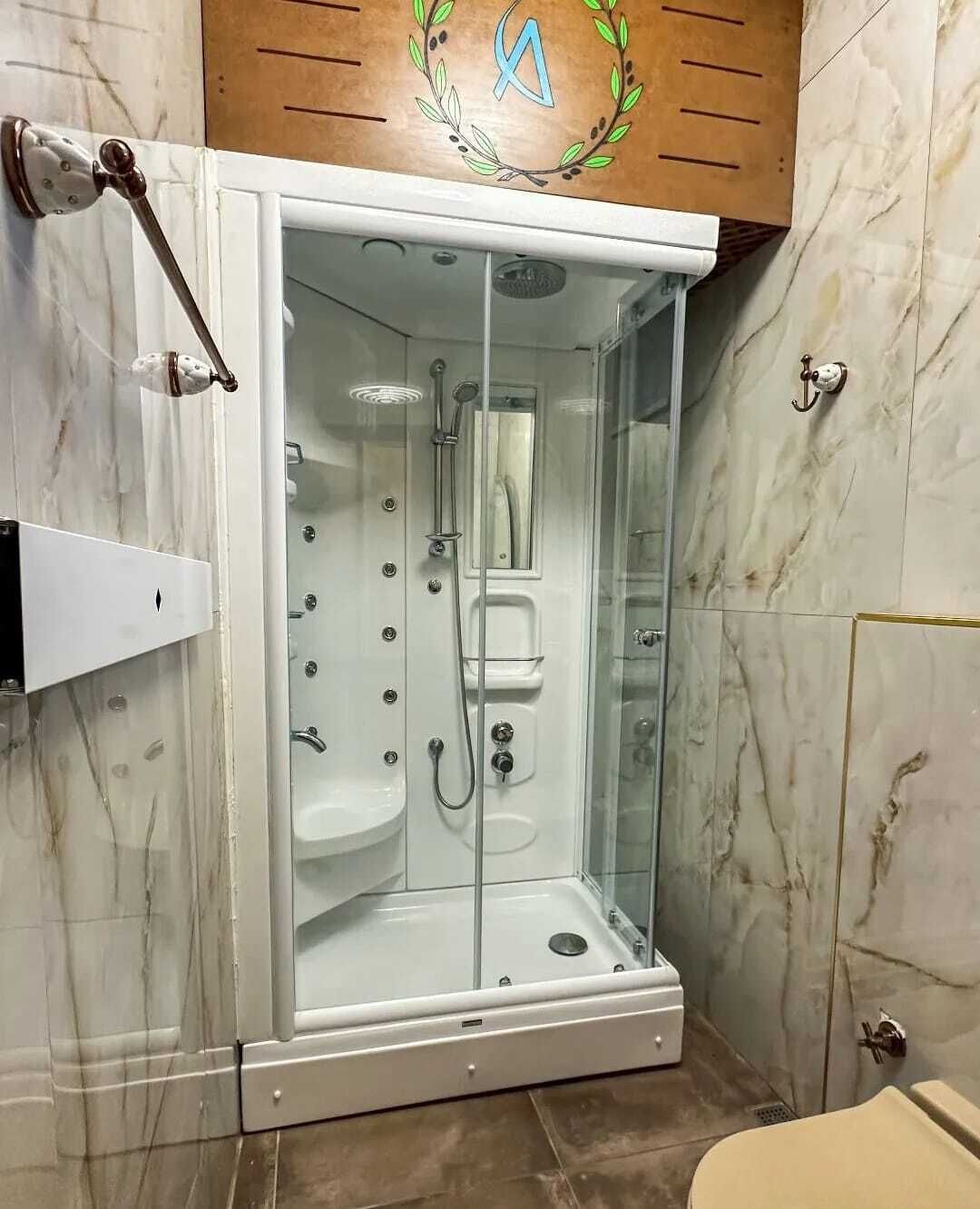 A glass-enclosed walk-in shower with a marble-tiled wall, a wooden panel above, and a white shelf unit to the left.