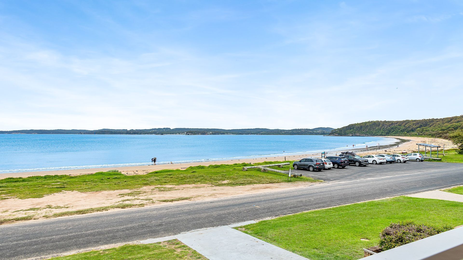 A coastal road with a beach on the right, cars parked along the side, and a clear view of the sea.