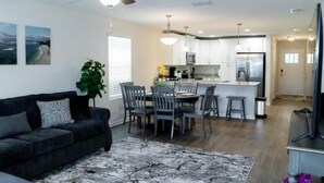 A modern living room with a black sofa, a dining area with a table and chairs, a kitchen with white cabinets, and a ceiling fan. - Near Airport & Beach Fully Fenced House (Jacksonville)