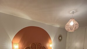 A bedroom with a curved headboard, a bed with a multicolored blanket, two bedside lamps, a small round table, and a wicker chair. - Gîte du Pommier 🍎 (Legé)