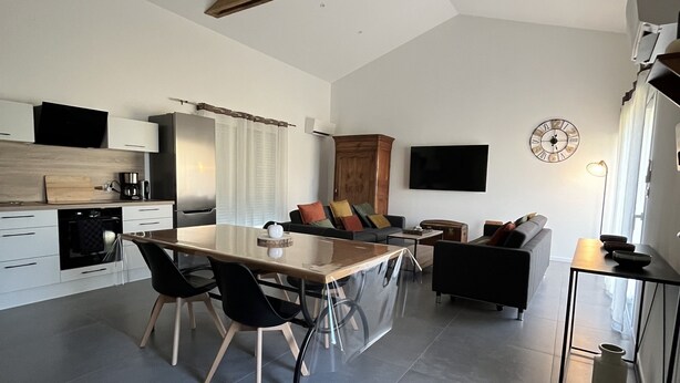 A modern kitchen with a central island, a dining area with chairs, a sofa, and a TV mounted on the wall. - Gîte du Pommier 🍎 (Legé)