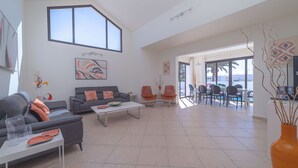 Un salon spacieux avec un canapé gris, une table basse blanche et des fauteuils orange. On aperçoit un coin repas avec une table et des chaises à travers une porte ouverte.