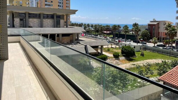 A balcony with a glass railing overlooking a landscaped area with palm trees and a view of the sea. - Vollga Apartment 9 (Durrës)