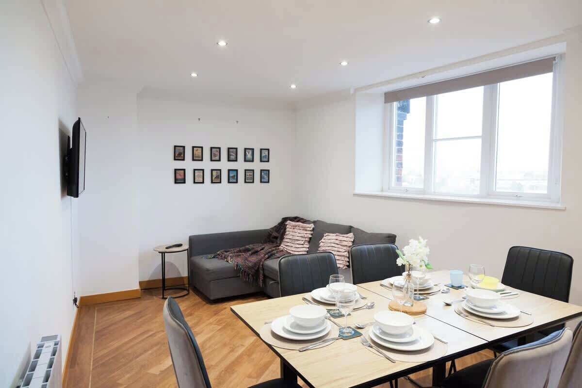 Un comedor con una mesa preparada para cuatro, un sofá gris y una ventana con persianas.