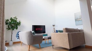 A modern living room with a flat-screen TV displaying 'NETFLIX', a grey armchair, a beige sofa, a small table, and a potted plant.