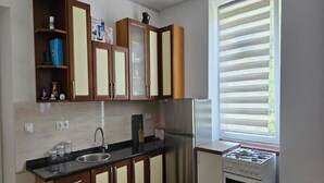 A compact kitchen with wooden cabinets, a stainless steel refrigerator, a gas stove, and a window with blinds.