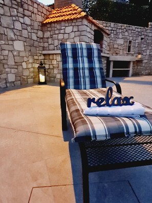 Una silla de lounge con un cojín a rayas y un almohadón que dice 'relax', ubicada en un patio con un muro de piedra y un farol al fondo.