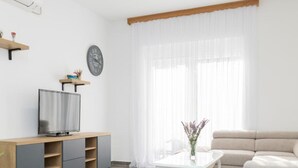 A modern living room with a flat-screen TV on a wooden TV stand, a beige sofa, a white coffee table, and a large window with sheer curtains.