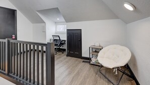 Una habitación con techo inclinado, una puerta negra, una silla blanca, una mesita y una ventana.