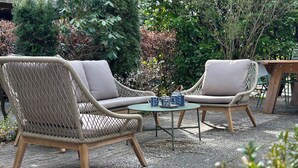 Two wicker chairs with cushions, a small round table with a few items on it, and a wooden bench in a garden setting.