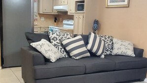 Un salon avec un canapé gris, des coussins à motifs, un fauteuil noir et un coin cuisine doté de meubles en bois et d’un réfrigérateur.