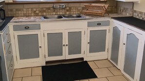 Una cocina con armarios blancos, encimera negra, ventana con persianas y salpicadero de azulejos.