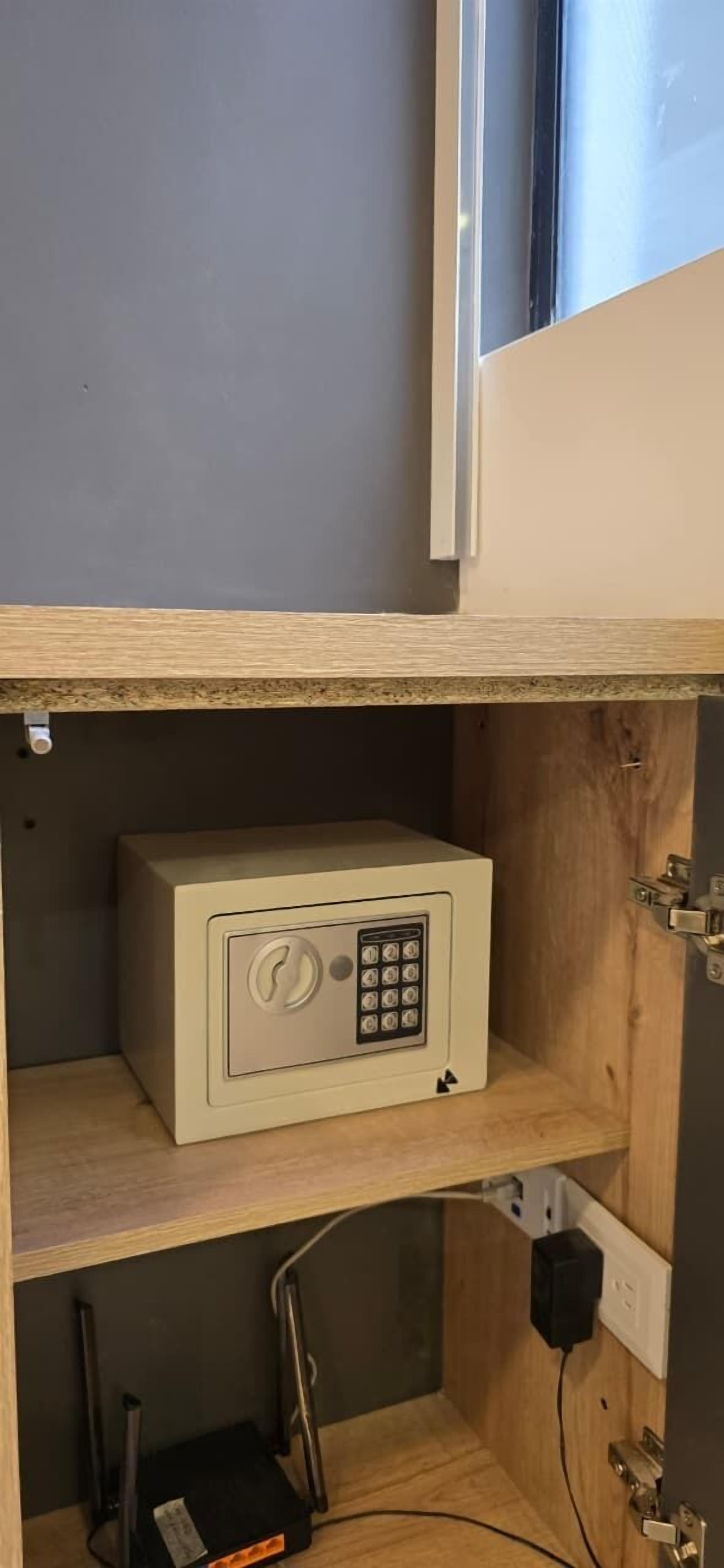 Una caja fuerte pequeña y beige sobre un estante de madera en una habitación con pared gris y una ventana.