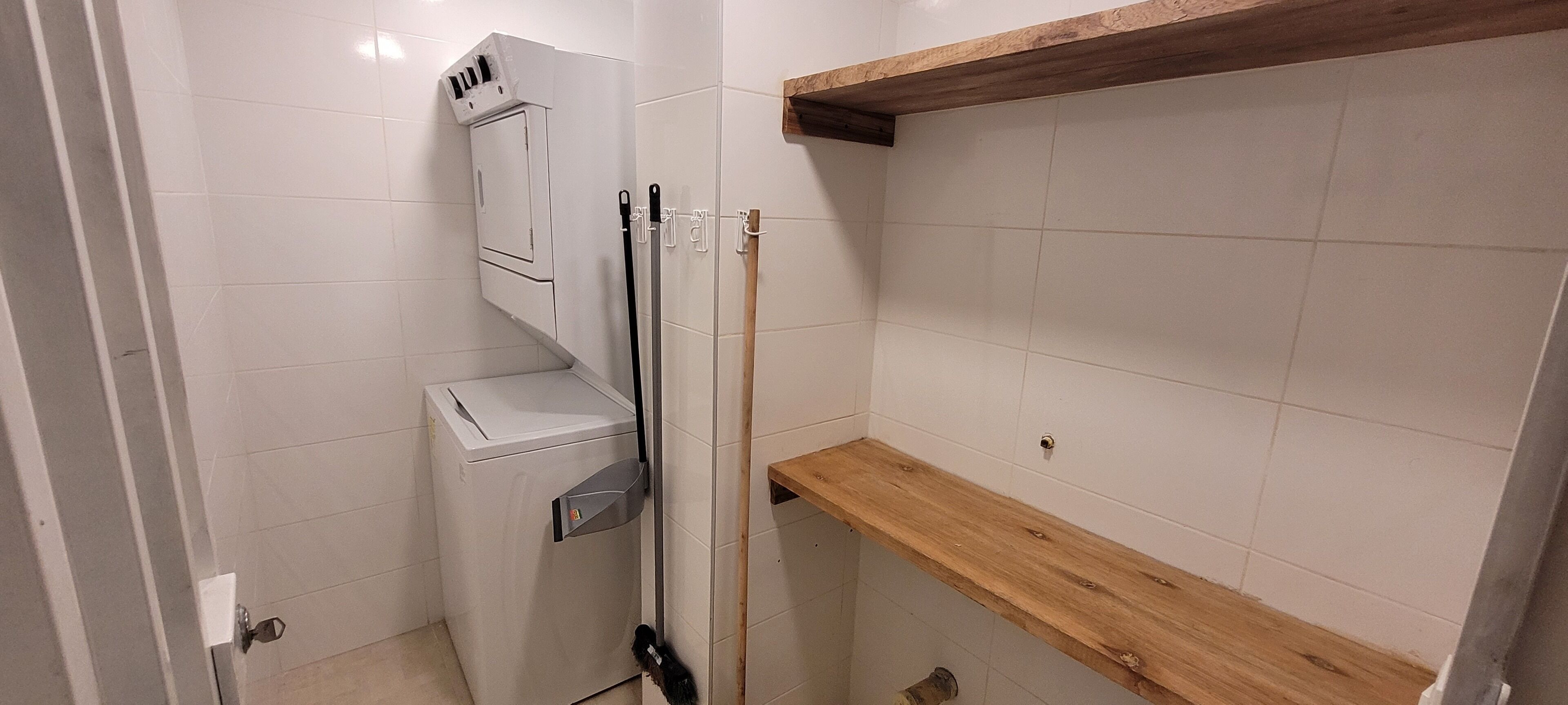 A compact bathroom with white tiles, a washing machine, a broom, and a wooden shelf.