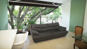 A living room with a grey sofa, a white chair, and a glass table. There is a balcony with a view of trees and a railing. - EDIFICIO FENICIA (Medellín)