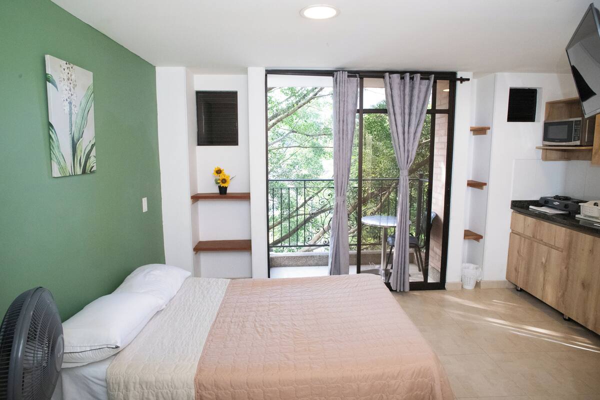 A bedroom with a bed, a fan, a green wall, a window with curtains, and a balcony with a chair.