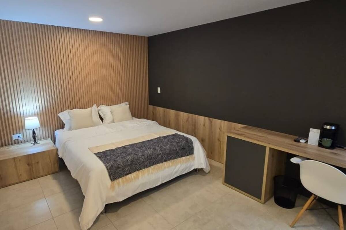 A bedroom with a bed, bedside table, lamp, desk, chair, and a small trash bin.