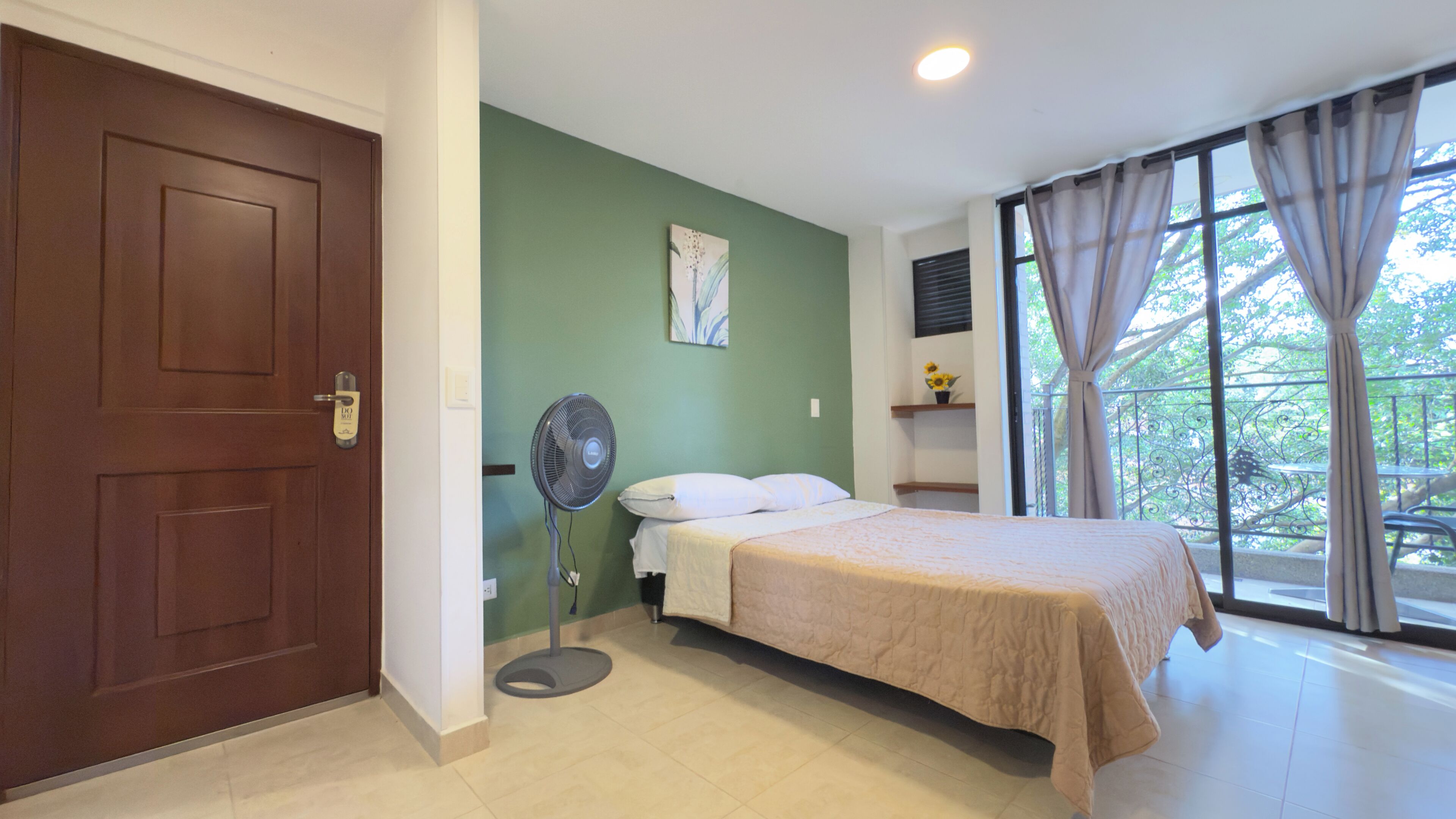 A bedroom with a bed, a standing fan, a door, a window with curtains, and a green wall.