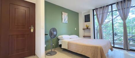 A bedroom with a bed, a standing fan, a door, a window with curtains, and a green wall.