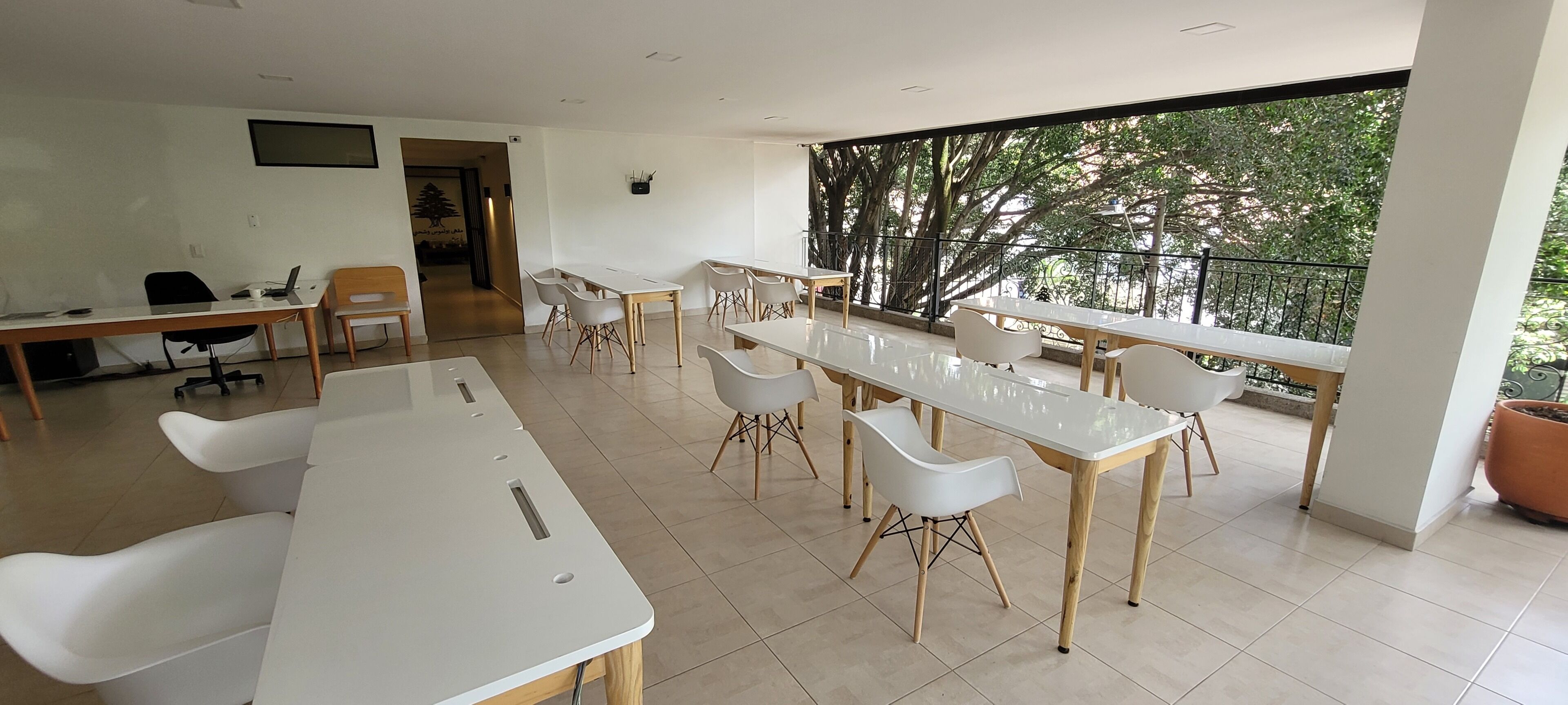 Une salle de conférence moderne, dotée de tables et de chaises blanches, d’une grande fenêtre donnant sur un paysage arboré et d’une porte menant à une autre pièce.