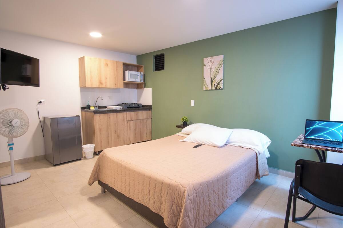 A compact room with a bed, a small kitchenette, a fan, and a laptop on a table.