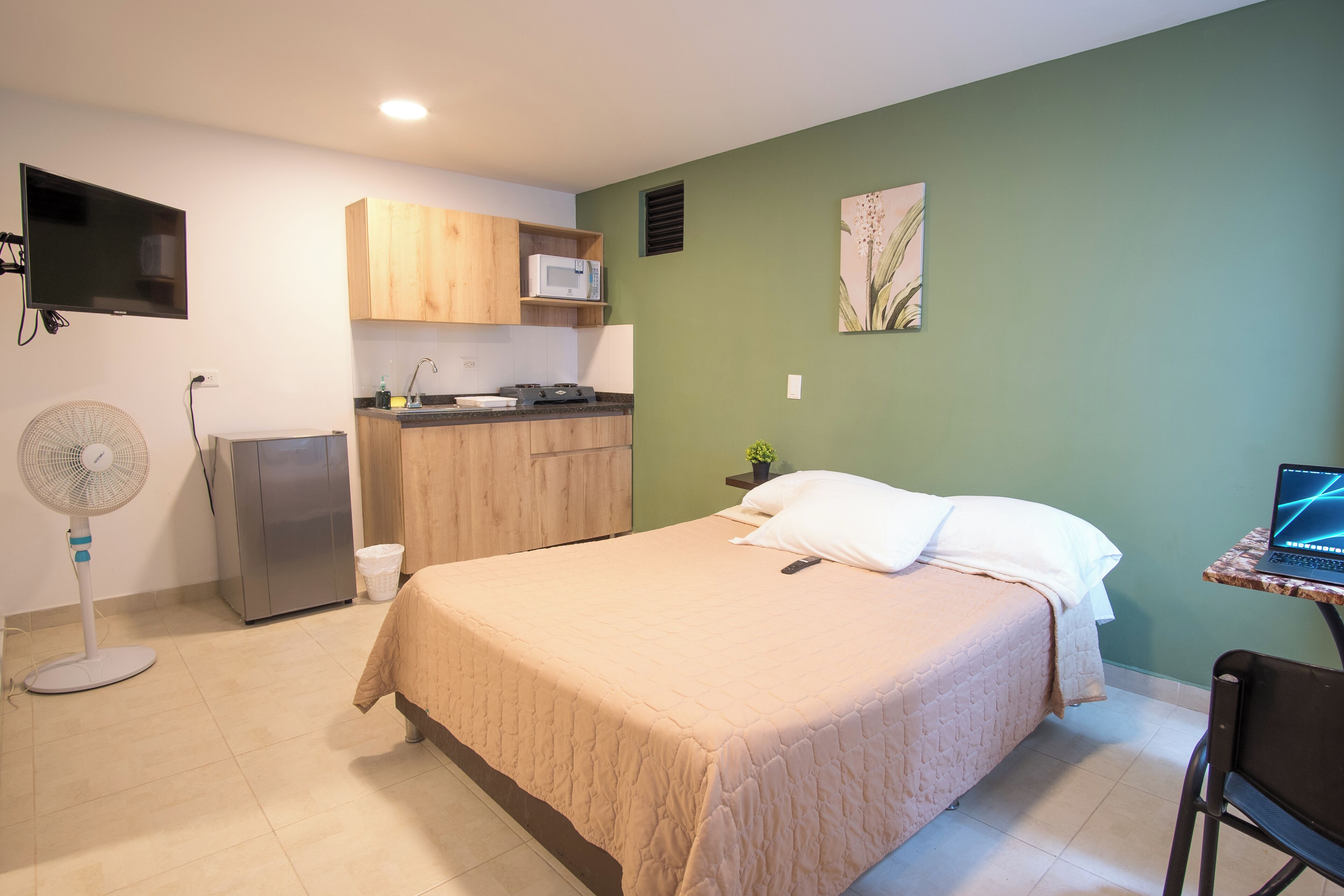 A compact bedroom with a bed, a small kitchenette, a fan, and a TV mounted on the wall.