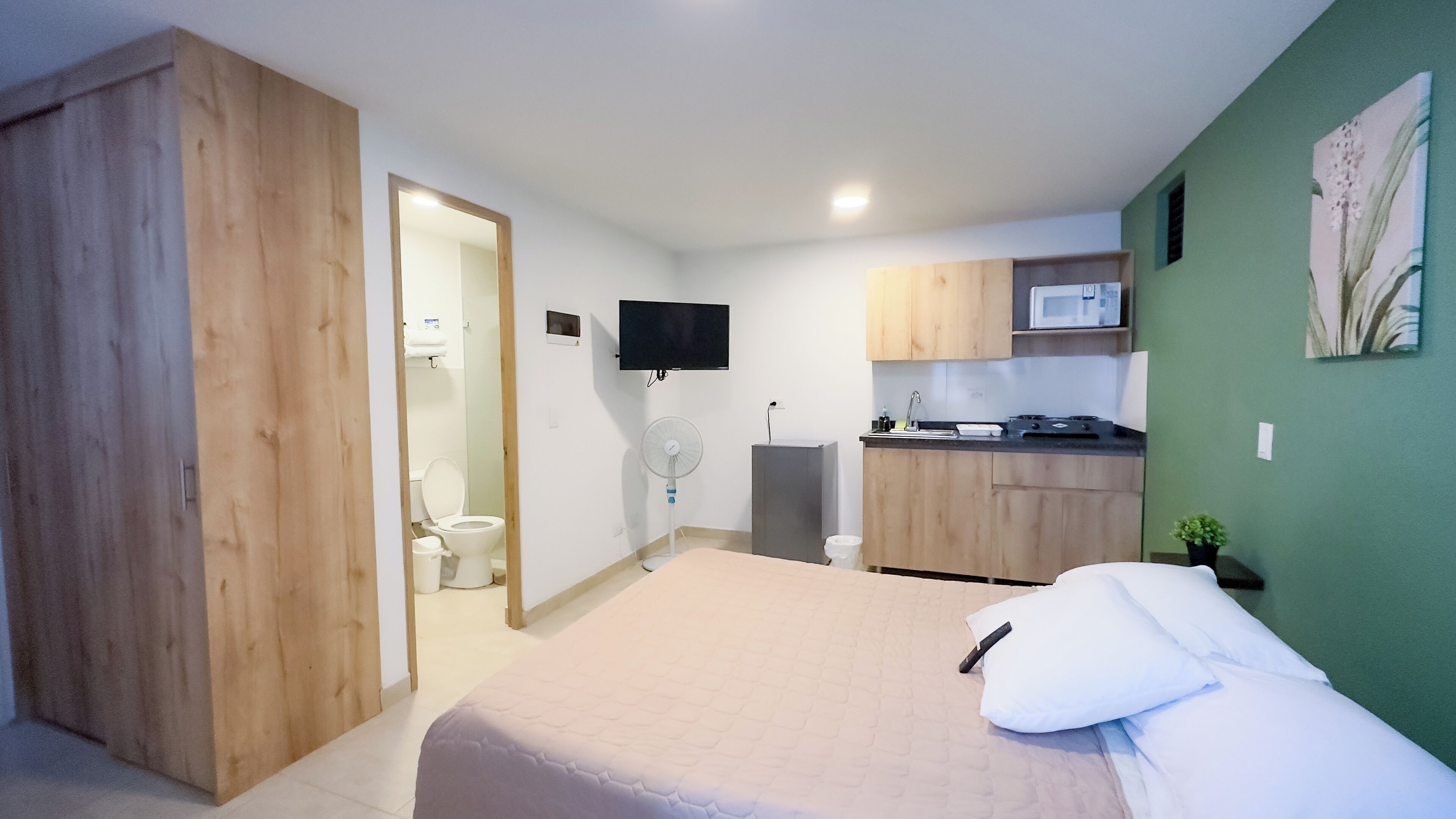 Une petite chambre comprenant un lit, une kitchenette, des toilettes et un téléviseur fixé au mur.