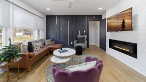 A modern living room with a fireplace, a ceiling fan, and a large window. - Chic and cozy home near the hospital: 3 comfy rooms, TVs, stylish amenities. (Gastonia)