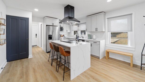 A modern kitchen with white cabinets, a central island with bar stools, a black range hood, and a large window. - Chic and cozy home near the hospital: 3 comfy rooms, TVs, stylish amenities. (Gastonia)