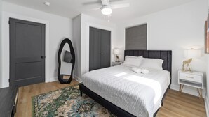 A bedroom with a bed, a ceiling fan, a nightstand, a mirror, and a door. - Chic and cozy home near the hospital: 3 comfy rooms, TVs, stylish amenities. (Gastonia)