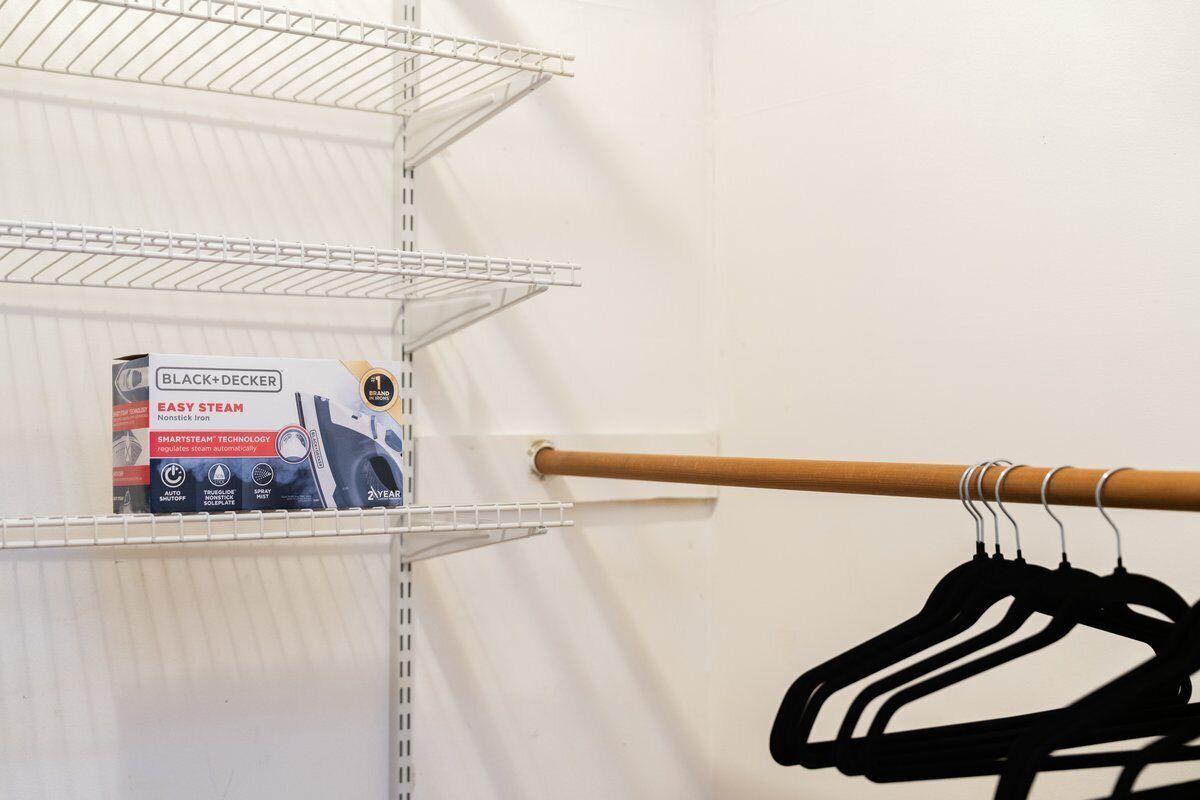 Ein Schrank mit einer Metallregaleinheit, einer Kiste mit der Aufschrift "BLACK+DECKER EASY STEAM" und einer Holzkleiderstange mit Kleiderbügeln.