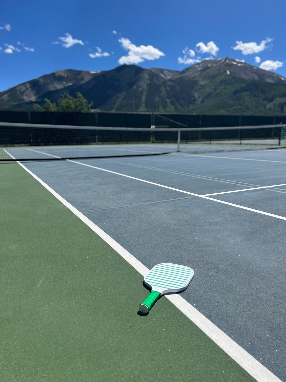 A tennis court with a green and white tennis racket lying on the ground, surrounded by a fence and mountains in the background.