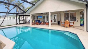 An outdoor swimming pool with a covered patio area, furnished with wicker chairs and a sofa, and a view of palm trees. - Ruby's Retreat, heated pool, private oasis, fenced backyard (Cape Coral)