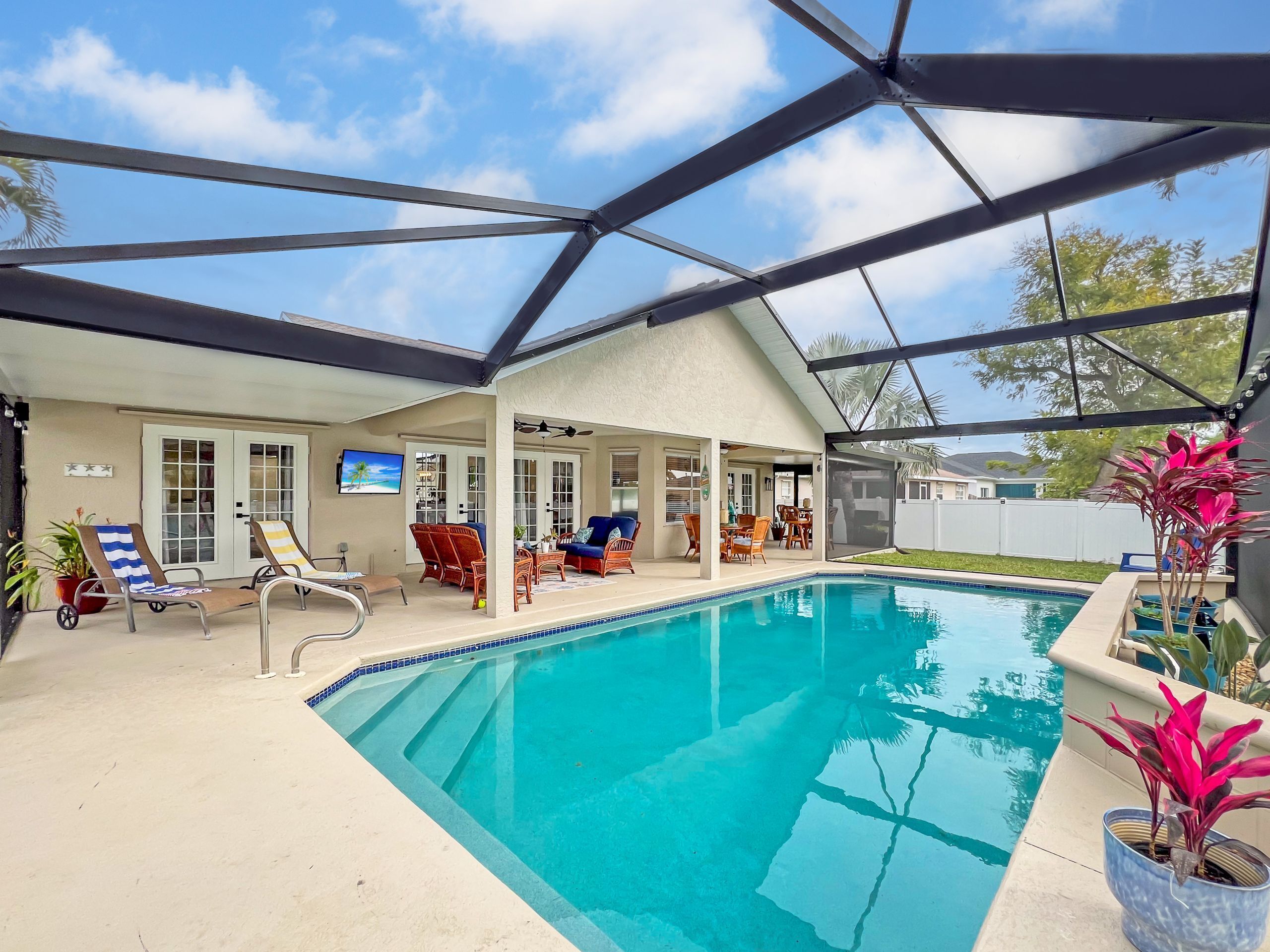 A covered outdoor swimming pool area with lounge chairs, a television, and a patio with furniture.