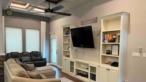 A living room with a beige sectional sofa, a small round ottoman, a flat-screen TV mounted on a white entertainment unit, and a ceiling fan. - Main House Listing 3BD/2BA

 (Edinburg)