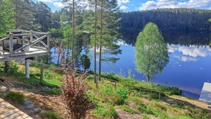 A lakeside cabin with a wooden deck, surrounded by dense forest and a clear view of the lake.