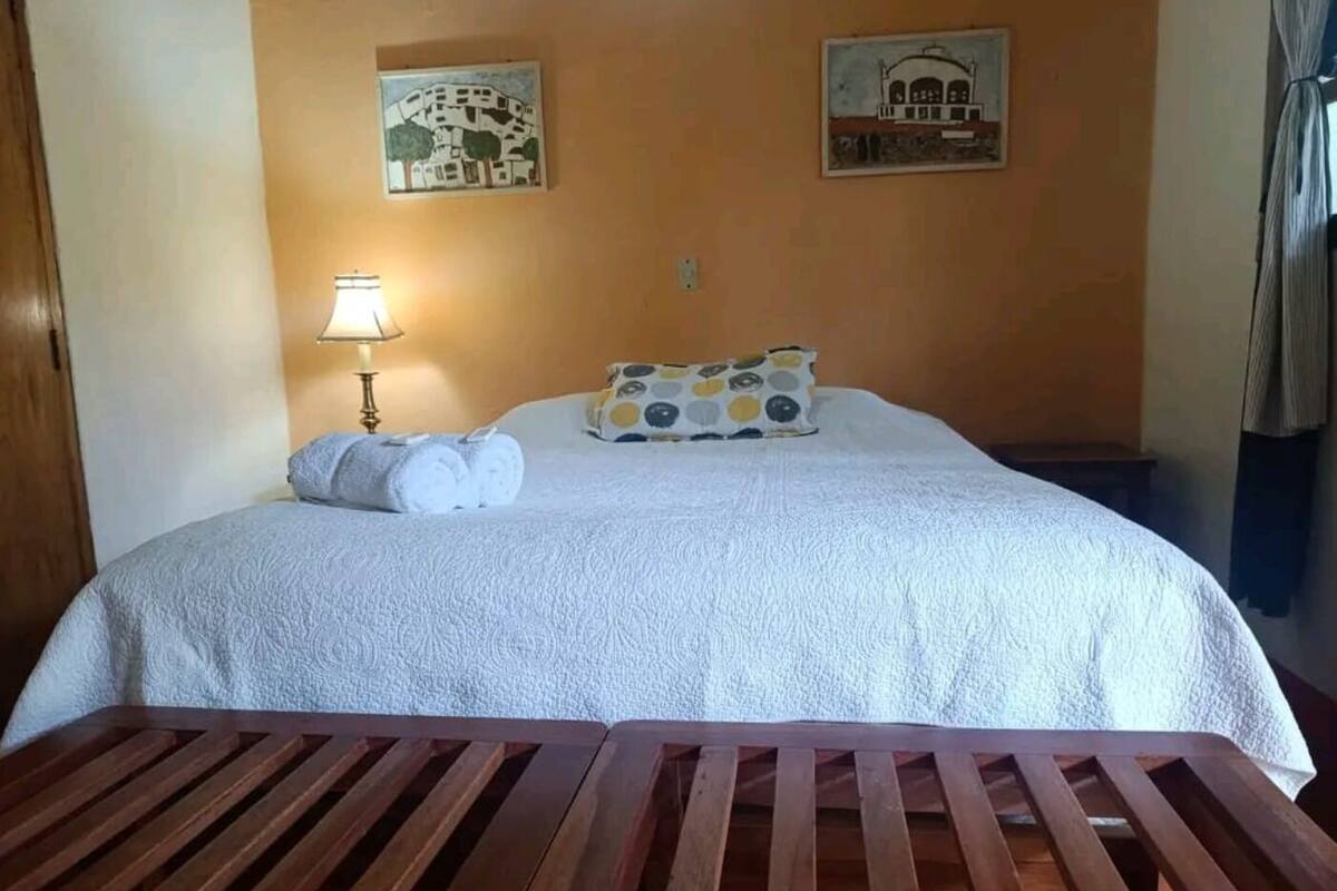 A hotel room with a bed, a wooden bench, two wall-mounted paintings, a lamp, and a patterned pillow.