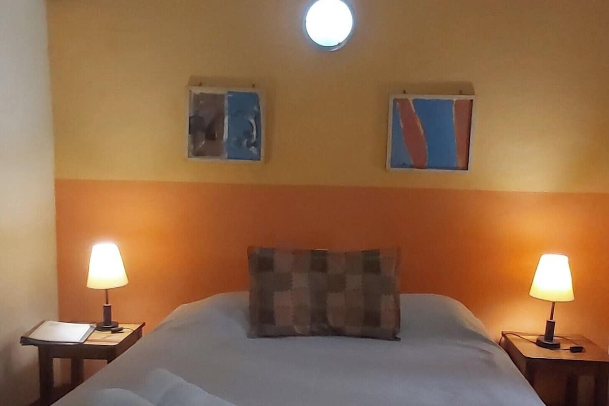 A hotel room with a bed, two bedside lamps, a ceiling light, and two wall-mounted artworks.