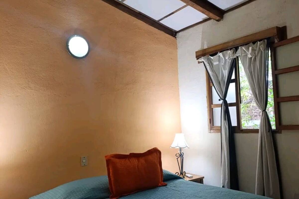 A bedroom with a bed, a ceiling fan, a window with curtains, and a wall-mounted lamp.