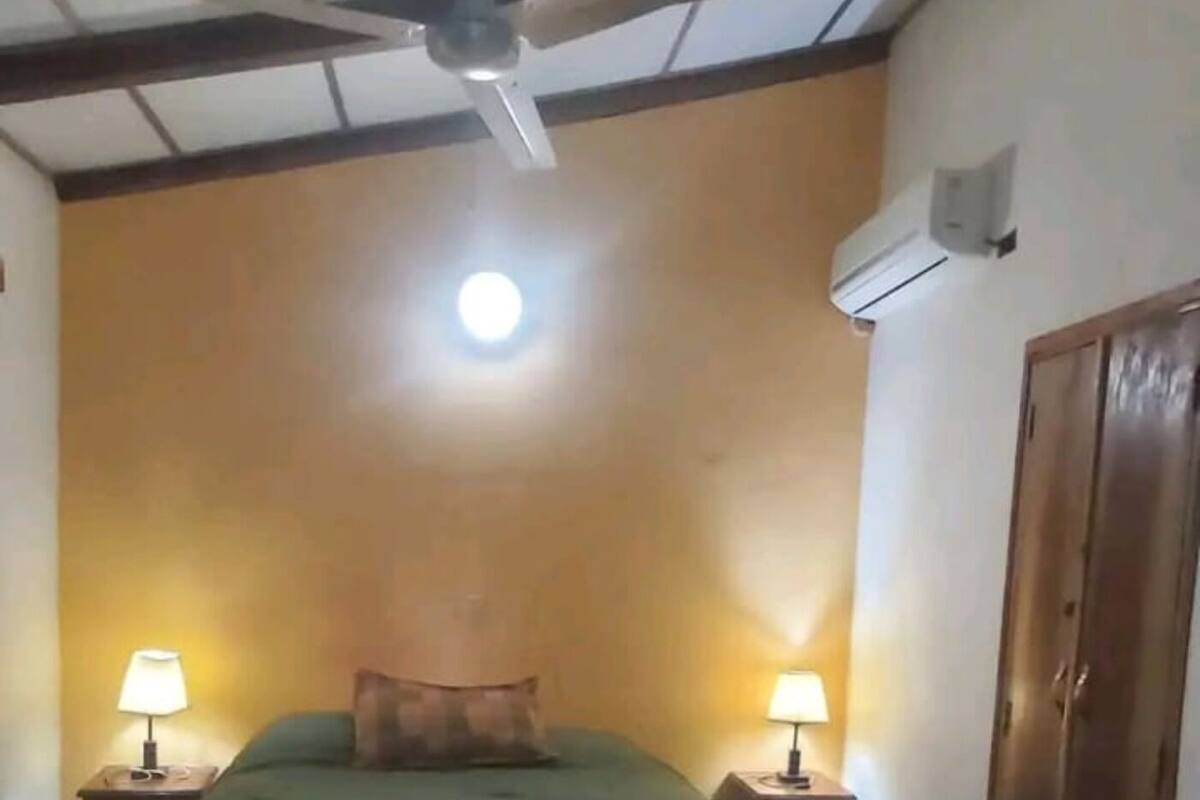 A room with a bed, two bedside lamps, a ceiling fan, an air conditioning unit, and a wooden wardrobe.