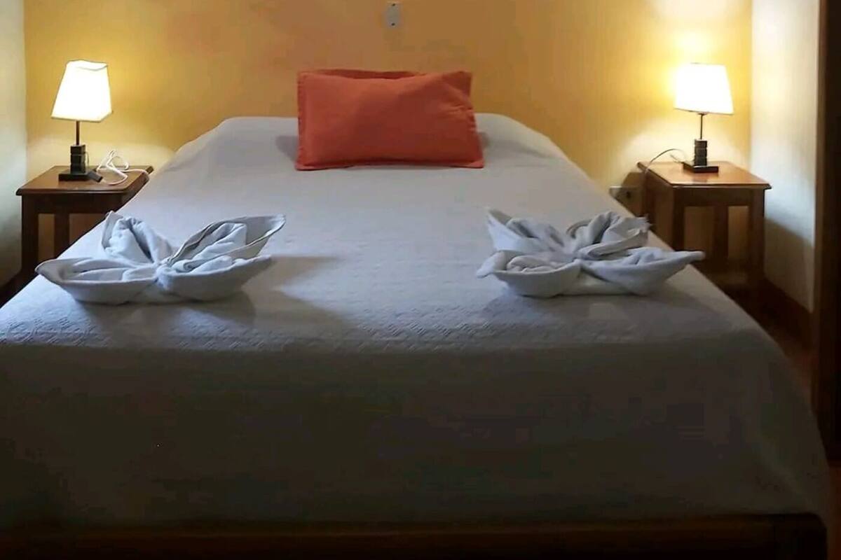 A hotel room with a bed, a single orange pillow, two bedside lamps, and an air conditioning unit.