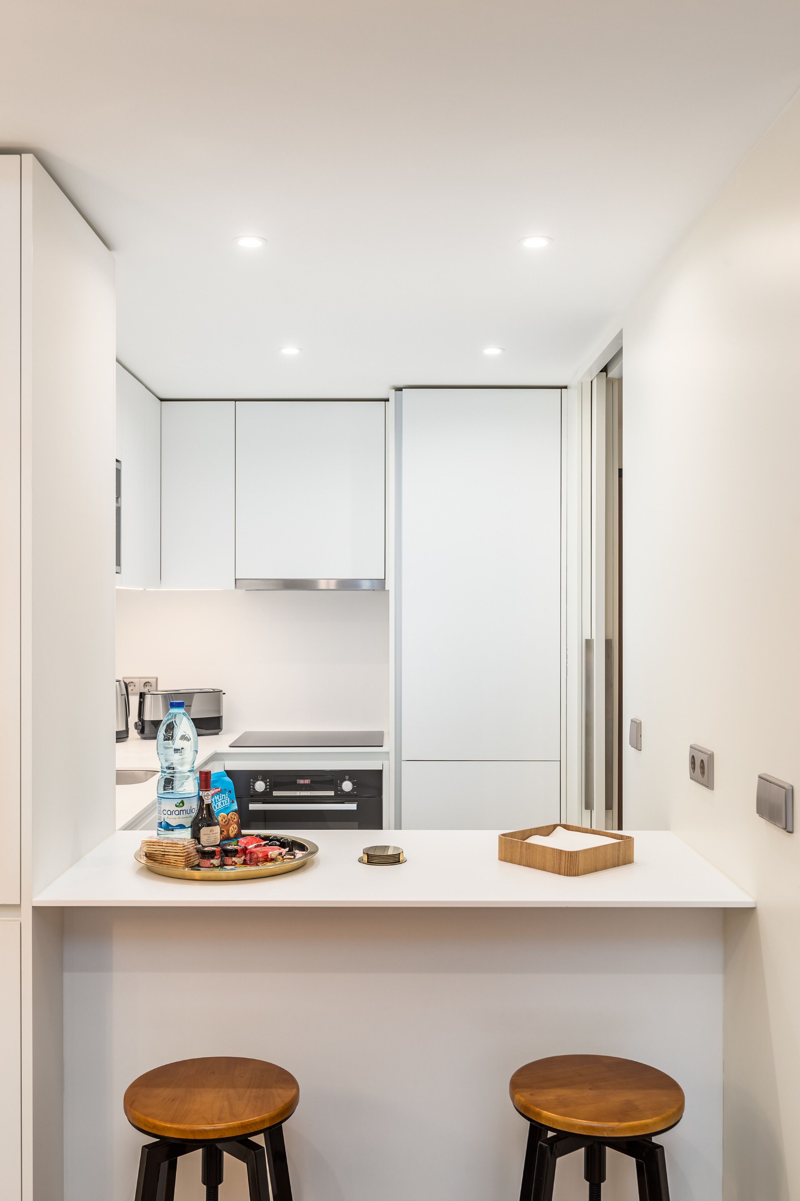 A compact kitchen with white cabinetry, a countertop, two wooden stools, and a breakfast tray with food and drinks.