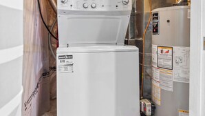 A compact laundry room with a stacked washer and dryer, visible pipes, and a basket of cleaning supplies.